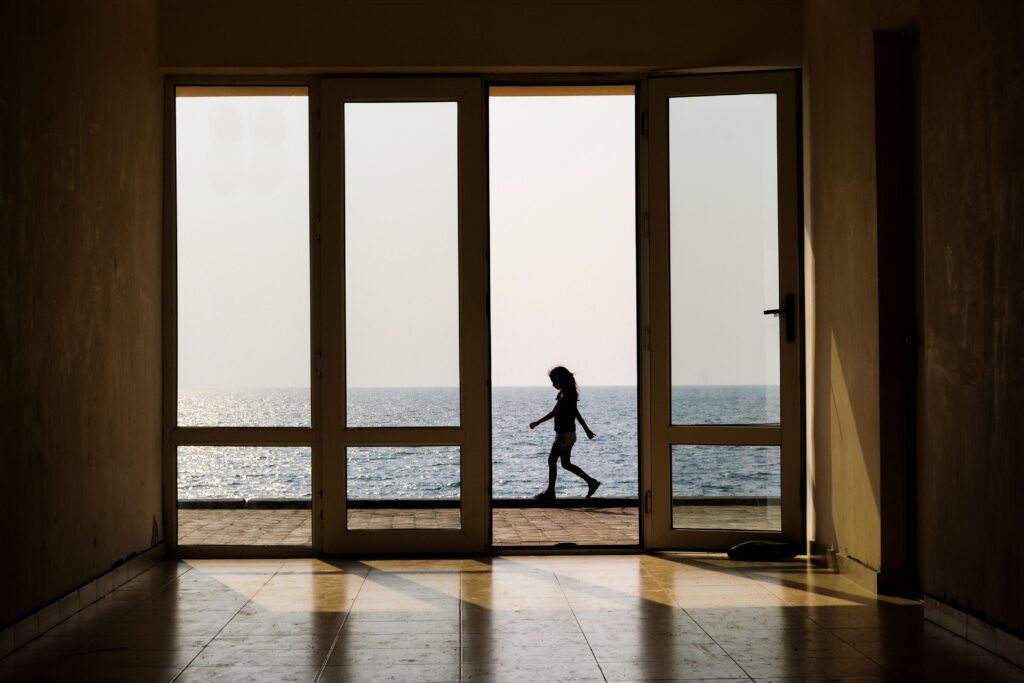 Through doors of building side view of silhouette of anonymous kid walking along embankment near sea on sunny day