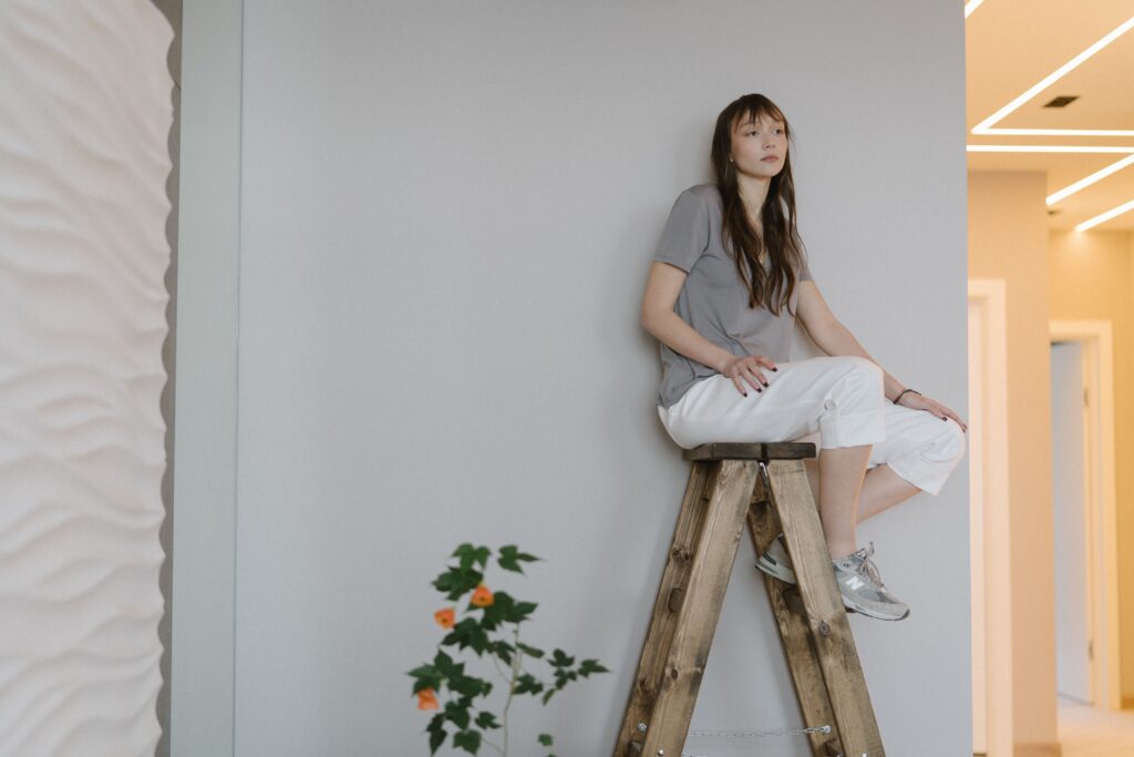A woman sits on a wooden ladder against a textured wall indoors, conveying calmness.